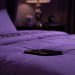Smartphone posé sur un lit, éclairage violet tamisé et ambiance nocturne intime dans une chambre élégante.
